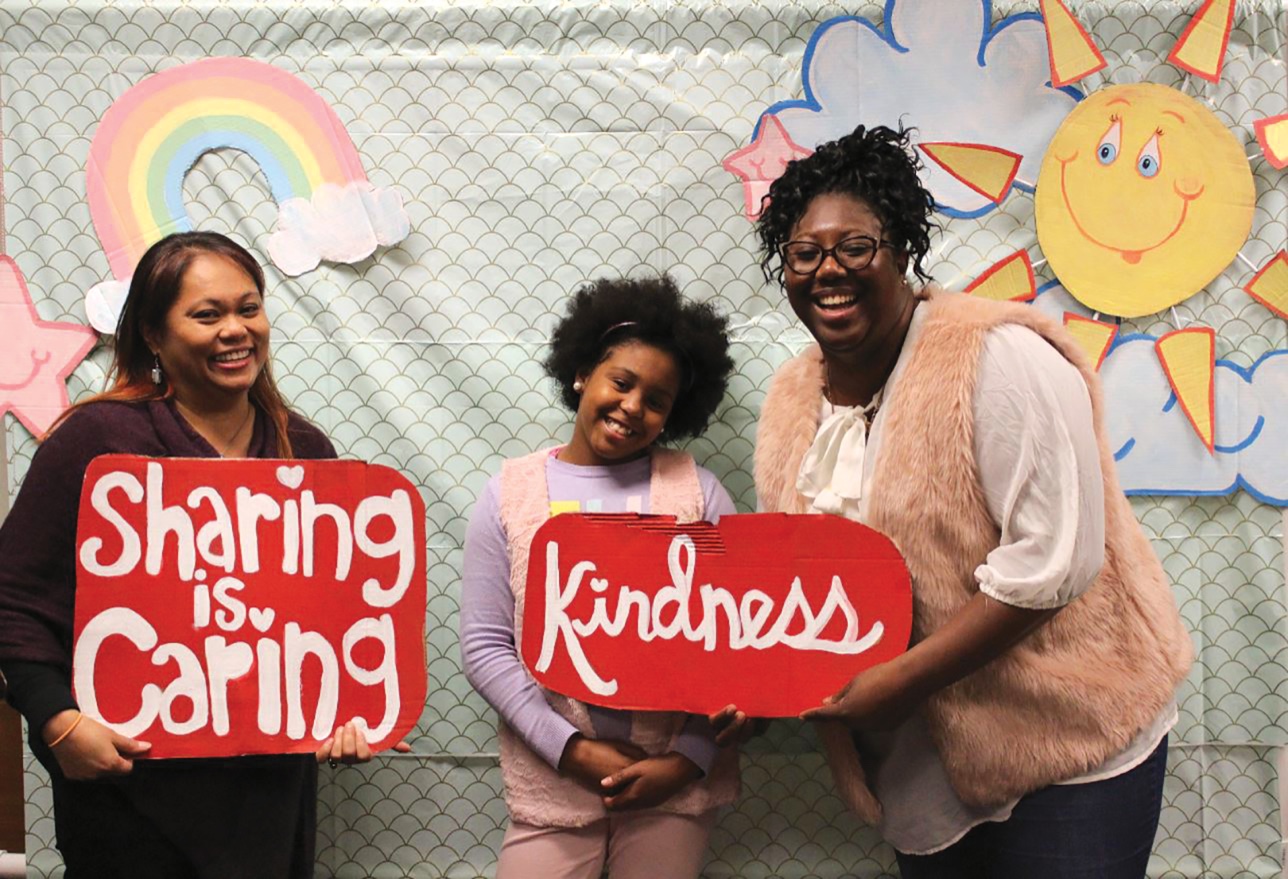 Lai-Lani Ovalles, Alaia Greer, and Amanda Greer hold handmade signs reading “sharing is caring” and “kindness”