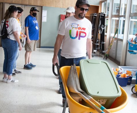 A man pushes a wheelbarrow with a white Team Up shirt on