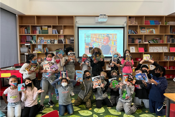 A group of masked kids hold books up in front of shelves lined with more books.