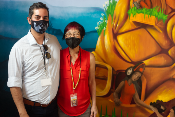 Ann Takamoto and Oscar Arana poser in front of a mural depicting a pastoral Native village 