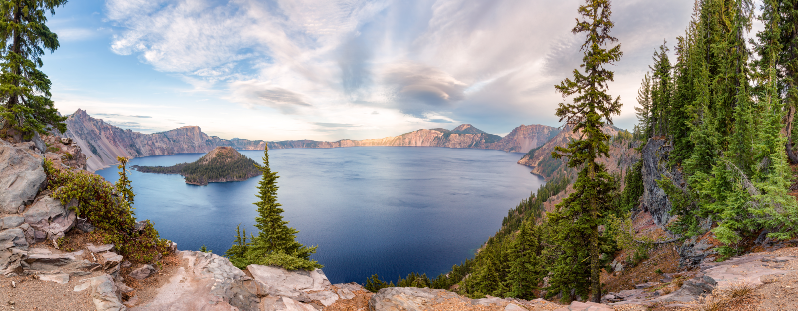 A wide photo of Crater Lake with clouds in the background and evergreens in the forefront