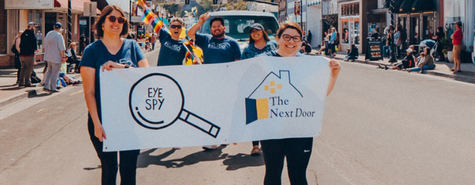 A group of people in a parade hold a banner saying The Next Door.