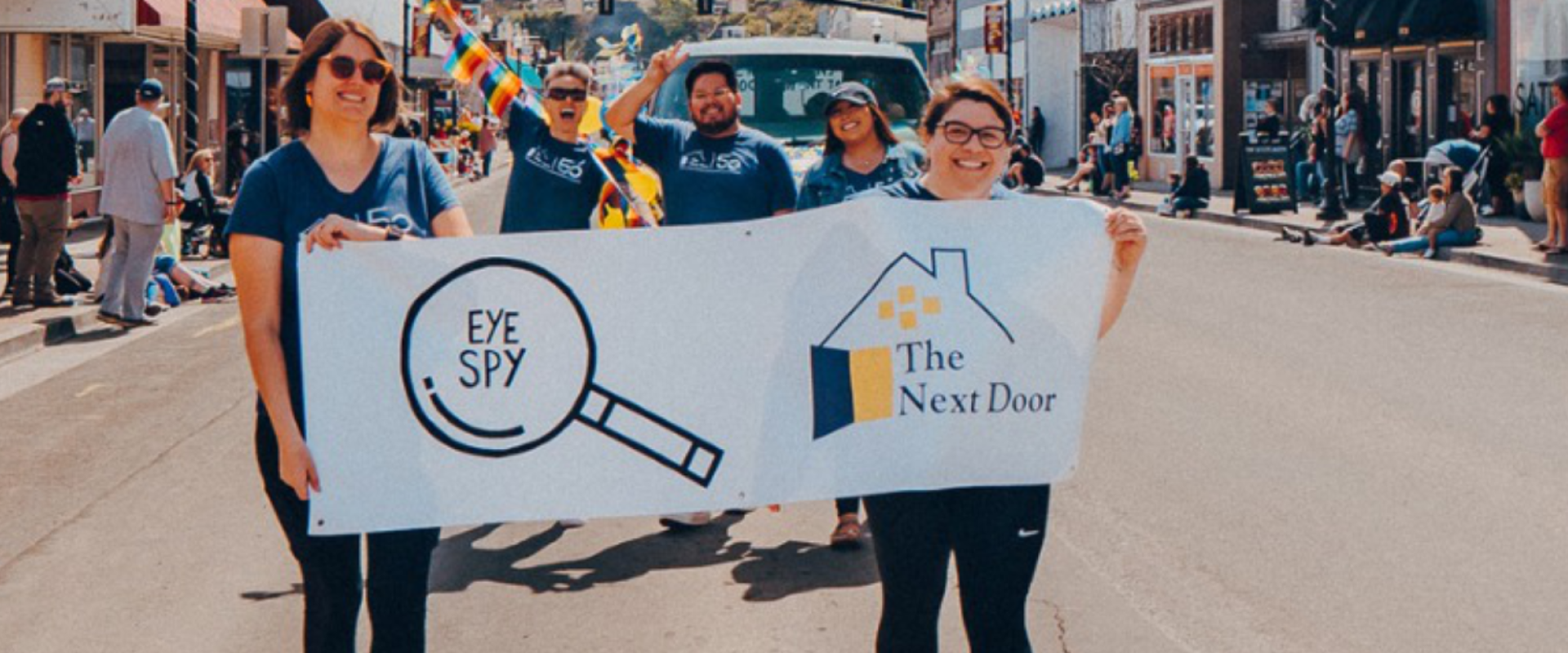 A group of people in a parade hold a banner saying The Next Door.
