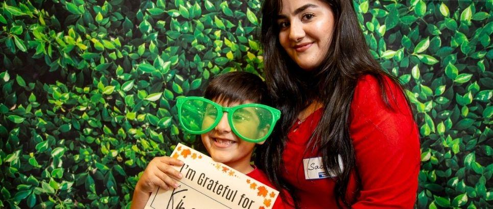 A mother and her child smile in front of a backdrop of greenery. The child is wearing oversize sunglasses and holding a sign that says I'm grateful for kindness