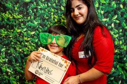 A mother and her child smile in front of a backdrop of greenery. The child is wearing oversize sunglasses and holding a sign that says I'm grateful for kindness