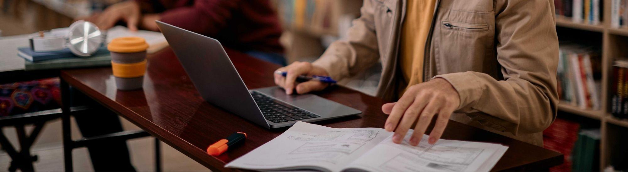 a hand touches paper and a laptop on a busy desk. Bookshelves are in the background out of focus.