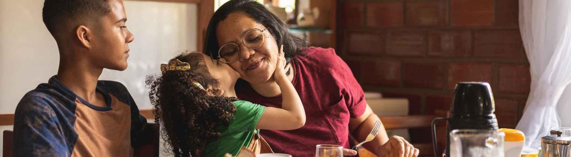 Mom with son and daughter eating breakfast at the table. The daughter is kissing mom on the cheek and mom is smiling with her eyes closed. The son is looking at them to the right.